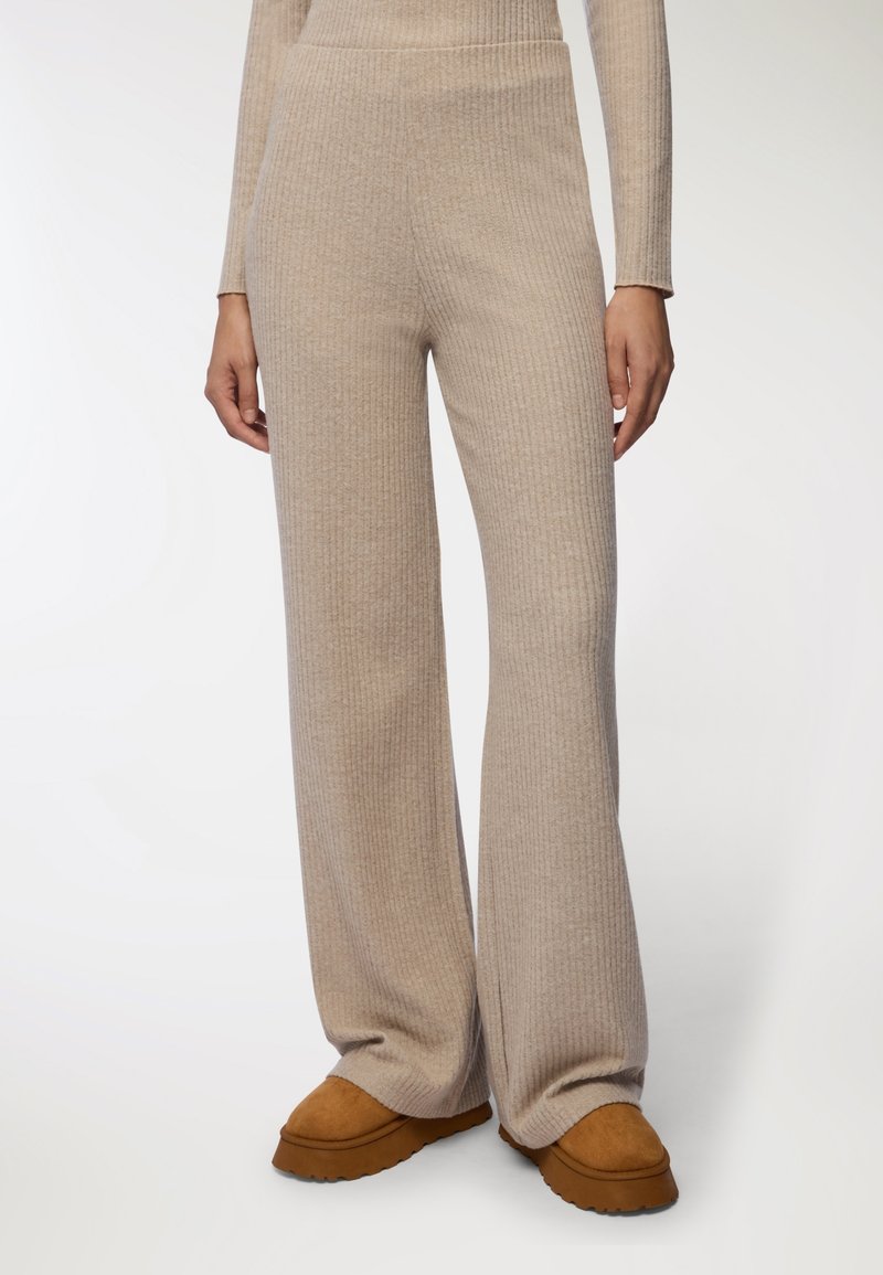 Beżowe, prążkowane spodnie dzianinowe z wysokim stanem i szerokimi nogawkami, wykonane z miękkiego, teksturowanego materiału. Stylizowane z brązowymi butami wsuwanymi.