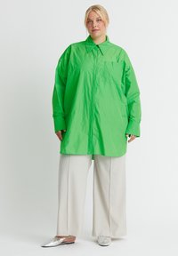 Lange mouwen, oversized shirt in felgroen, gemaakt van lichte stof, met een knoopsluiting aan de voorkant en een borstzak, gecombineerd met beigekleurige wijde broek.