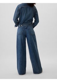 Conjunto de chaqueta de mezclilla y jeans de pierna ancha en azul medio. La chaqueta presenta un diseño estructurado con bolsillos y detalles de botones. Los jeans tienen un corte holgado.
