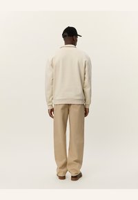 Sudadera de cuello redondo beige con puños y base acanalados, combinada con pantalones anchos color beige claro y una gorra negra, vista desde la parte trasera.
