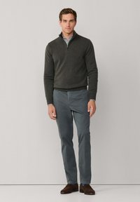 Donkergrijze trui met rits over een lichtblauw en wit gestreept shirt, gecombineerd met grijze broek en bruine schoenen, staand tegen een neutrale achtergrond.