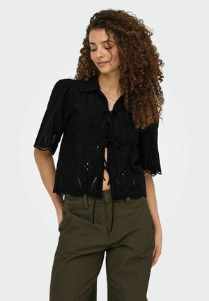 Jeune femme aux cheveux bouclés porte un chemisier noir à œillets avec des liens devant et un pantalon vert olive, debout les mains dans les poches.