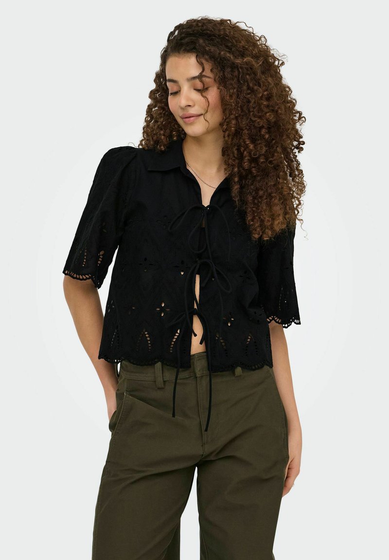 Jeune femme aux cheveux bouclés porte un chemisier noir à œillets avec des liens devant et un pantalon vert olive, debout les mains dans les poches.