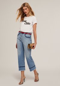 Camiseta blanca de manga corta con "Très Chic" en letras negras, combinada con unos vaqueros cortos azul claro y un cinturón rojo, sosteniendo un bolso de mano dorado.