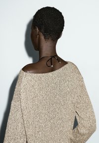 Persoon met kort, krullend haar, gekleed in een beige gebreid vest dat één schouder blootlegt en een zwarte knoopketting aan de achterkant van de nek.