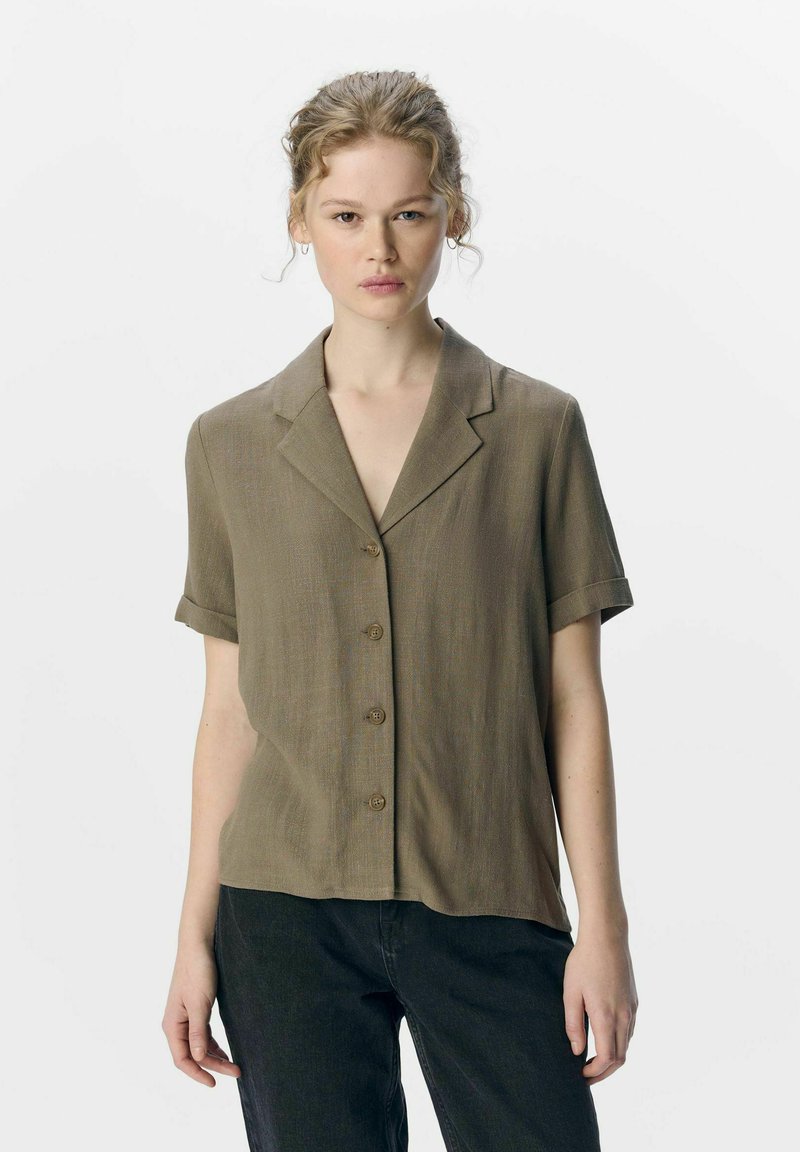 Camisa de manga corta con botones en verde oliva, con un cuello con muesca y una textura suave, combinada con pantalones oscuros.