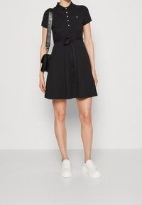Czarna sukienka polo z krótkimi rękawami, zapięciem na guziki, paskiem w talii i rozszerzanym dołem. Połączona z białymi trampkami i czarną torbą.
