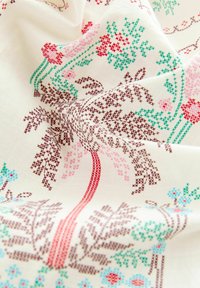 Witte stof met ingewikkelde bloemembroderieën in rood, roze, groen en blauw. Patronen omvatten bomen en bloemen met verschillende texturen.