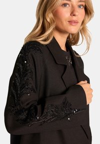 Chaqueta negra con un diseño estructurado, que presenta una tela texturizada y elaborados embellishments de lentejuelas en un patrón de hojas en las mangas.