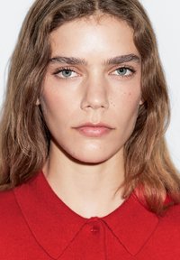 Nahaufnahme eines Gesichts mit langen, welligen braunen Haaren, klarer Haut und hellblau-grünen Augen. Trägt ein leuchtend rotes Hemd mit Kragen und Knöpfen.