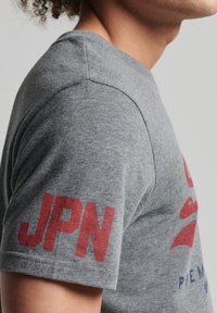 T-shirt en coton gris à manches courtes. Présente un texte "JPN" rouge sur la manche gauche et un design graphique multicolore à l'avant. Texture douce.