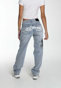 A person stands wearing a white cropped shirt and light blue baggy jeans with "Ed Hardy" logo and graphic design on the back pocket.