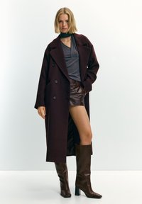 Femme portant un long manteau foncé par-dessus un haut gris, un short en cuir marron et des bottes marron mi-mollet, debout devant un fond uni.