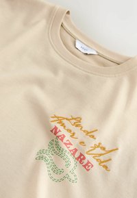 T-shirt beige à col rond avec texte brodé « Lindo seja amor e fada NAZARÉ » et motif abstrait vert cousu sous le texte.