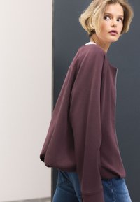 Pārāk liela bordo krāsas džemperis ar fakturētu audumu, platiem piedurknēm un rāvējslēdzēja aizdari. Valkāts kopā ar gaiši zilām džinsa biksēm uz neitrālas fona.