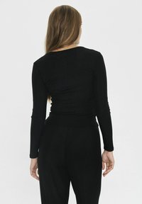 Woman with long brown hair wearing a fitted black long-sleeve top and black pants, standing facing a plain white background.
