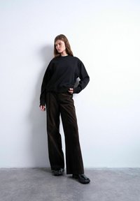 Femme en sweat-shirt noir, pantalons larges foncés et chaussures noires, debout contre un mur blanc sur un sol gris.