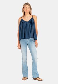 Granatowa top bez rękawów z marszczonym materiałem, zestawiony z jasnoniebieskimi dżinsami w fasonie dzwonów i brązowymi sandałami. Widoczne proste akcenty biżuteryjne.