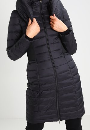 Vrouw draagt een lange, zwart doorgestikte pufferjas met capuchon, handen die de kraag aanpassen, rits aan de voorkant gesloten.