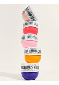 Stapel aus fünf runden Körpercremebehältern in den Farbtönen Rosa, Rot, Orange und Lila, jeweils mit dekorativen weißen Etiketten, auf denen die Produktnamen stehen.