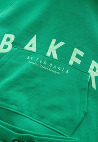 Vihreä huppari, jossa on suuret valkoiset kirjaimet, jotka kirjoittavat "BAKER" ja teksti "BY TED BAKER, Lontoo, Yhdistynyt kuningaskunta." Pehmeä, teksturoitu kangas.