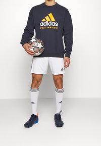 Sudadera azul marino con el logo "adidas REAL MADRID" en amarillo y blanco, pantalones cortos blancos, calcetas blancas con franjas negras y zapatos de fútbol azul marino. Sostenido un balón de fútbol.