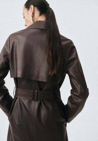 Brązowy skórzany płaszcz trench z paskiem w talii, szerokim kołnierzem oraz warstwowym tyłem. Gładka faktura i dopasowany krój.