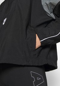 Svart vindtålig jacka med grå detaljer, zip-ficka, elastiska ärmslut och vit pipning. Har Nike-loggan på bröstet.