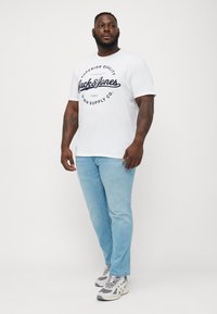Camiseta blanca con estampado azul marino, jeans de color azul claro y zapatillas grises. La camiseta presenta un logo circular. El modelo está de pie en un fondo neutral.