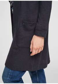 Lång, mörkgrå cardigan med en ficka och svarta knapppdetaljer, texturerat tyg, bärs över blå jeans. Ärmfickorna är fållade.
