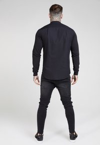 Maglietta nera a maniche lunghe con una texture liscia, design aderente e orlo arrotondato, abbinata a jeans scuri e sneakers nere.