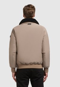 Homme portant une veste matelassée beige avec un col en fourrure noire et des poignets côtes, vu de dos contre un fond uni.