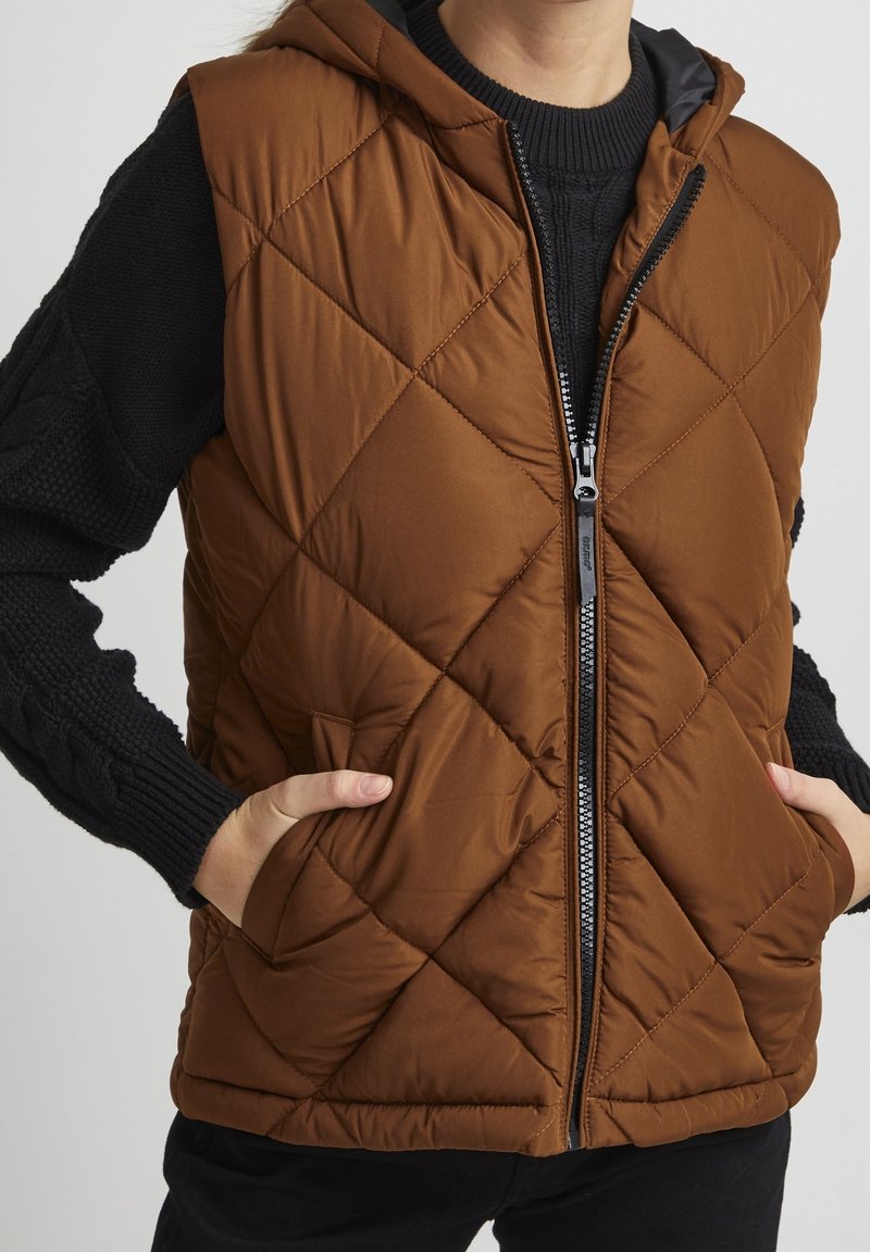Personne portant une veste sans manches matelassée marron avec une capuche et un pull en tricot noir à manches longues, les mains dans les poches de la veste.