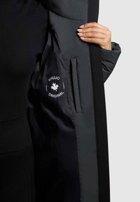 Čierny vystužený kabát má na vnútornej strane hladkú tmavú látku s bielou logom a emblémom javorového listu, vrátane bočnej vrecka.