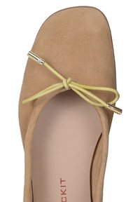 Chausson plat en suède beige avec un orteil carré, équipé d'un lacet en ruban jaune et d'un détail en doré sur le nœud. Doublure intérieure lisse.