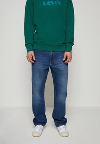 Sweat-shirt vert avec logo Levi's bleu, associé à un jean denim bleu. Des baskets blanches complètent le look, avec un design classique.