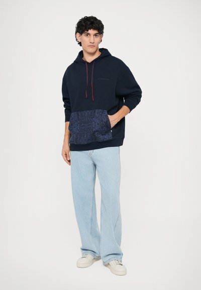 Marineblauer Hoodie mit roten Kordeln und gemustertem blauen Fronttasche, kombiniert mit hellblauen weit geschnittenen Jeans und weißen Turnschuhen.
