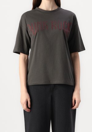 Henkilö pukeutuneena tummanharmaaseen oversized T-paitaan, jonka rinnassa lukee "PUNK ROCK" punaisilla niiteillä ja löysät mustat housut.