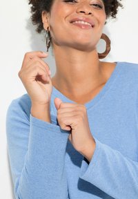 Maglietta a maniche lunghe di colore azzurro chiaro con scollatura a V, realizzata in tessuto morbido. Le mani del modello sono posizionate vicino al colletto, mostrando una vestibilità rilassata.