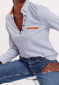 Button-up shirt with a blue and orange geometric pattern, featuring a brown collar accent and a chest pocket. Worn with blue denim jeans.
