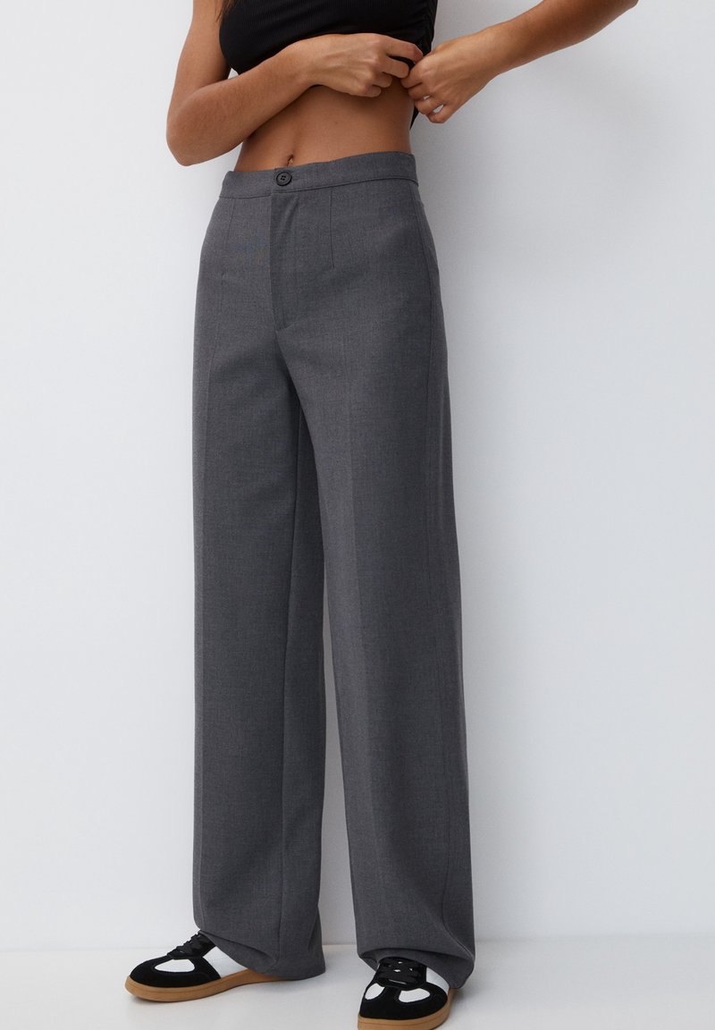 Personne portant un pantalon gris taille haute à jambes larges et des baskets noir et blanc, ajustant un top court noir contre un mur blanc.