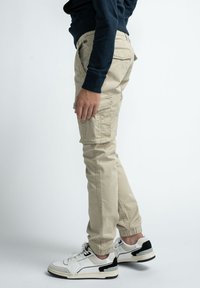 Pantalon cargo beige avec jambes fuselées, doté de poches latérales et de poignets élastiques, associé à des baskets blanches et noires.