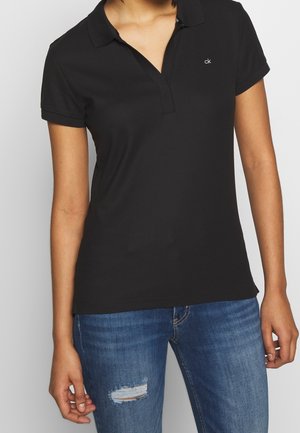 Czarna koszulka polo z kołnierzykiem, krótkimi rękawami i subtelnym detalem logo. Połączona z zniszczonymi niebieskimi dżinsami, które mają przetarcia.