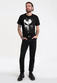 Black cotton t-shirt featuring a white graphic of a skull design, paired with black jeans and black lace-up boots, standing against a light background.