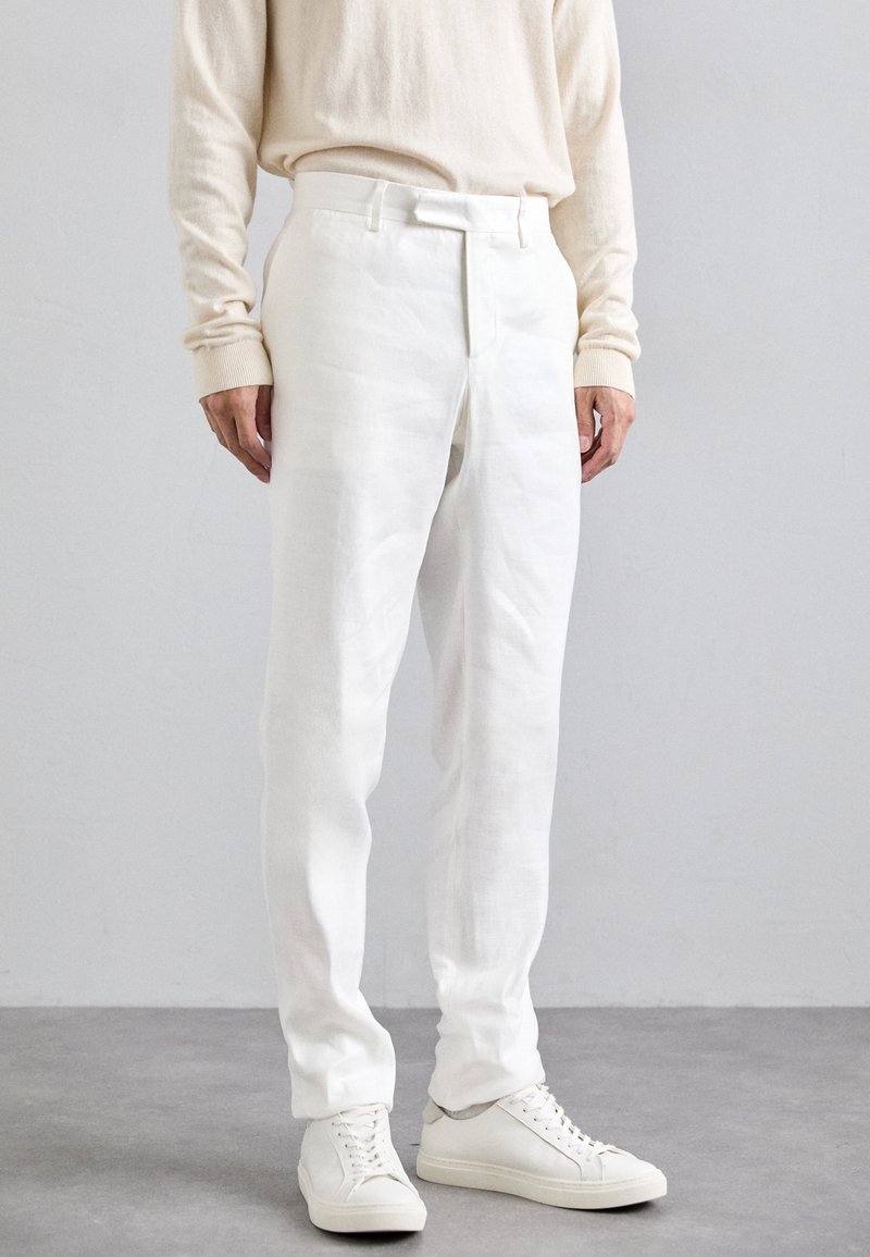 Hvide linned bukser med en tætsiddende pasform, sømløs foran lukning og sidelommer, parret med hvide sneakers. Stoffet er let og tekstureret.