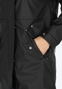 Svart vattentät jacka med en slät struktur, tryckknappar och sneda fickor. Har en hög krage och en stilren, minimalistisk design.