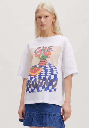 Jonge vrouw met lang blond haar draagt een wit oversized t-shirt met een kleurrijke bloem- en tafelafbeelding en een blauwe roezel denim rok.