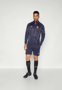 Vīrietis, kurš valkā tumši zilu Puma sporta jaku un šortus, melnas Puma zeķes un naglu apavus, stāv pret vienkāršu gaišu fonu.