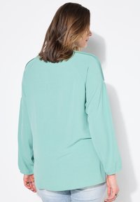 Maglietta verde menta a maniche lunghe con vestibilità ampia, dotata di maniche raglan e polsini elasticizzati. Tessuto morbido con una leggera lucentezza.