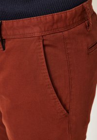 Rust-colored pants with a smooth texture, featuring a side pocket and subtle stitching details. Waistband has a ribbed knit design.